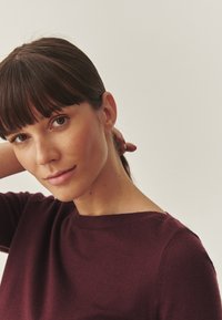 Burgunderfarbener Pullover mit Rundhalsausschnitt und langen Ärmeln, der eine weiche Textur aufweist. Das Model hat glattes braunes Haar und einen neutralen Hintergrund.
