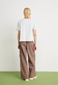 Une personne avec les cheveux blonds courts se tient sur un sol vert face à un mur blanc, portant un t-shirt gris clair, un pantalon cargo marron et des baskets crème.