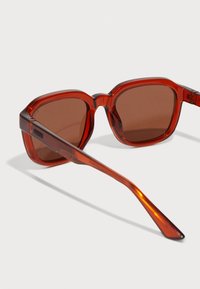 Gafas de sol rojas translúcidas con lentes marrones, montura cuadrada y brazos robustos. El diseño notable incluye bordes angulares y un acabado brillante.
