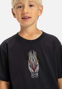 Camiseta de algodón negra con un gráfico de llamas en el centro en blanco, rojo y naranja, y un logo de DC debajo. Manga corta y cuello redondo.