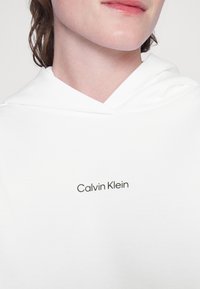 Sudadera con capucha blanca que presenta un cordón y un logo impreso de "Calvin Klein" en negro. Textura suave con un corte relajado y diseño minimalista.