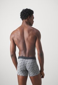 Graue Boxer-Shorts mit schwarzem elastischem Bund, das ein wiederholtes Logo-Textmuster aufweist. Modell von hinten vor einem hellen Hintergrund betrachtet.