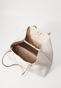 Sac à main beige matelassé en cuir lisse avec intérieur doublé. Il dispose d'une fermeture à pression, d'une poche zippée et d'un logo embossé, avec deux poignées pour l'épaule.