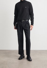 Černý svetr s kulatým výstřihem a malým emblémem, kombinovaný s černými padnoucími kalhotami a černými šněrovacími botami, v jedné ruce drží psaníčko.