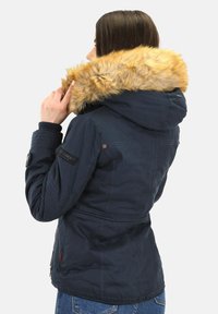 Vrouw in een marineblauwe jas met een capuchon met bontafwerking, die haar kraag aanpast, gezien van achteren, tegen een effen lichte achtergrond.