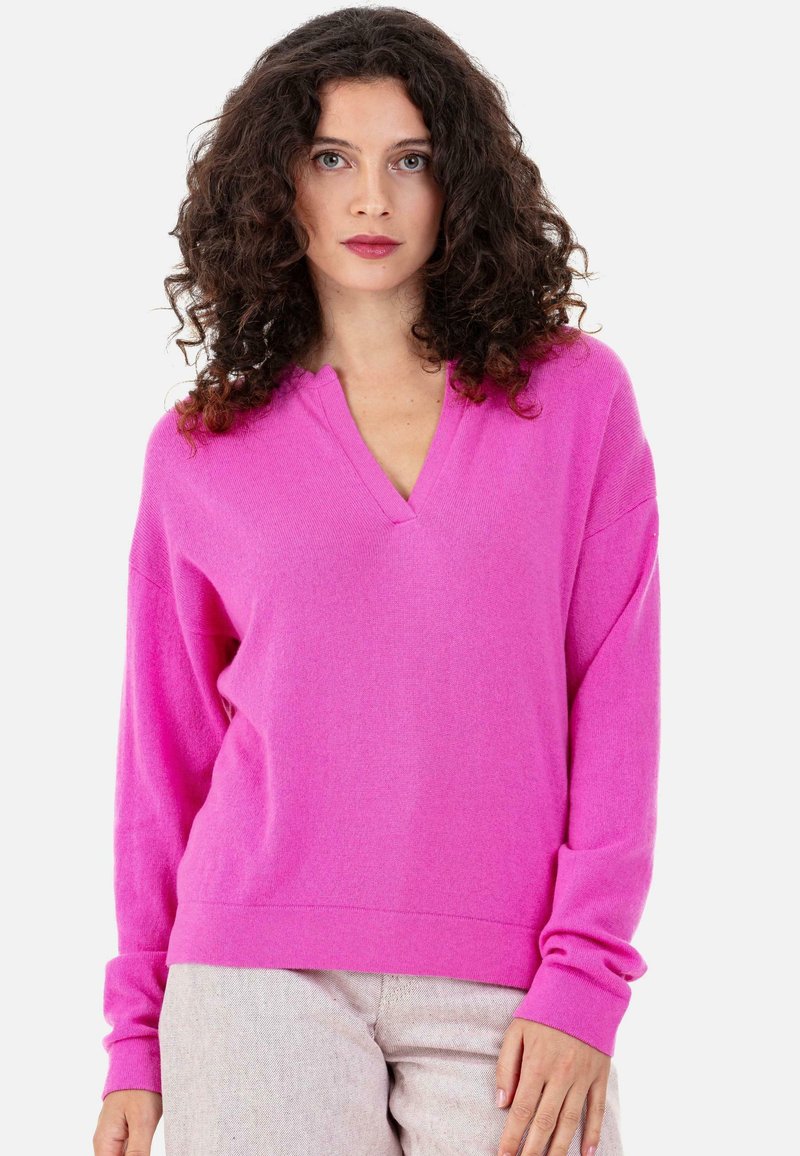 Pull en maille rose avec un col en V, coupe ample et manches longues. Texture douce et poignets côtelés, assorti à un pantalon de couleur claire.