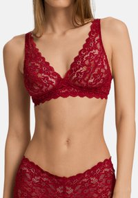 Červený krajkový bralet s květinovým vzorem, představující hluboký výstřih do V a vlnité okraje. Jemná, průsvitná textura s nastavitelnými ramínky.