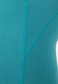 Zbliżenie na turkusową tkaninę z wyraźnymi pionowymi i skośnymi szwem na gładkiej powierzchni tekstylnej.