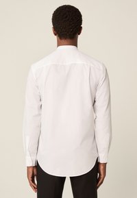 Bijela košulja s dugim rukavima, zakrivljenim rubom, kopčama na manžetama i klasičnim ovratnikom. Izrađena od glatkog materijala, s diskretnim detaljima prošivanja.