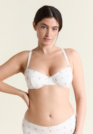 Mujer de cabello oscuro con un sujetador blanco de flores y ropa interior a juego, de pie con una mano en la cadera contra un fondo beige.