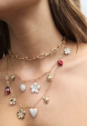 Nærbillede af en kvindes hals, der bærer lagdelte guldkæder med vedhæng, herunder hjerter, blomster, mariehøne, jordbær og en firkløver.