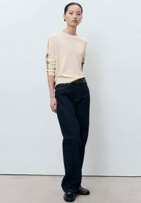 Femme jeune debout portant un haut à manches longues beige, un jean large noir, une ceinture noire, des mocassins noirs et des boucles d'oreilles en or sur un fond uni.