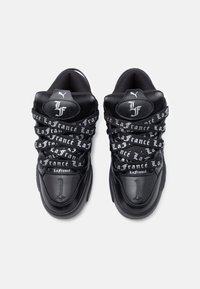 Ζευγάρι μαύρων αθλητικών παπουτσιών με χοντρές λευκές κορδόνες με το λογότυπο "La France" και λευκό λογότυπο στην αφρώδη γλώσσα, με θέα από πάνω σε λευκό φόντο.