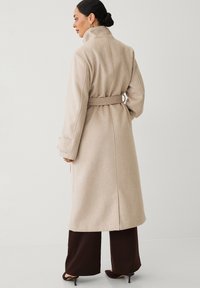Beige wollen jas met een hoge kraag, een ceintuur in de taille en lange mouwen. Heeft een strakke, op maat gemaakte silhouet en zichtbare stikseldetails.