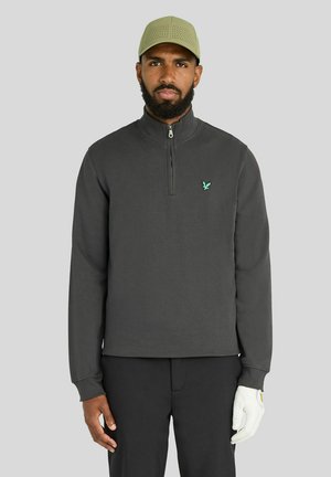 Mand iført en mørkegrå kvart lynlås-pullover, sorte bukser, lys grøn kasket og hvid golfhandske, stående mod en ensfarvet grå baggrund.