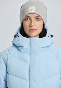Jasnoniebieska kurtka puchowa z wysokim kołnierzem, z widocznymi szwami i regulowanymi czarnymi sznurkami. Noszona z szarym, prążkowanym czapką beanie.