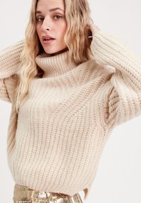 Pull en tricot beige avec un col haut et large et une texture côtelée, présentant des manches amples et une coupe décontractée. Jupe en sequins dorés brillants en dessous.