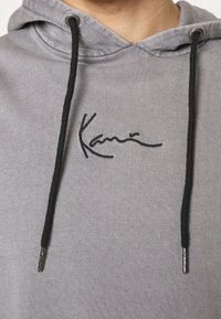 Sweat à capuche gris avec cordons noirs et un petit logo brodé en noir, style signature, centré sur la poitrine.