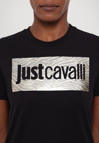 Svart t-skjorte med sølvgrafikk med "Just Cavalli" og et zebra-stripes mønster. Rund hals og korte ermer.