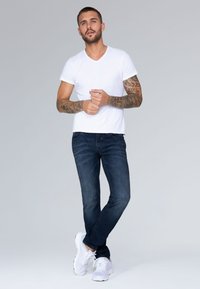 Camiseta blanca de cuello en V, jeans de color azul oscuro y zapatillas deportivas blancas. Diseño ajustado con mangas cortas y textura lisa, con tatuajes en los brazos.
