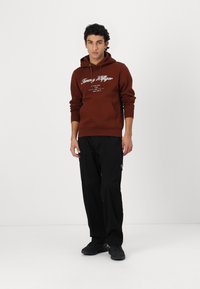 Brun hettegenser med hvit logo-tekst, lommer foran og snor i hetten; kombinert med svarte bukser og svarte joggesko. Enkel, uformell design.
