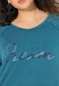 Tyrkysová košile s výstřihem ve tvaru lodi, s potiskem na přední straně, který zní "LETS Dream" v modrých a stříbrných kamíncích.