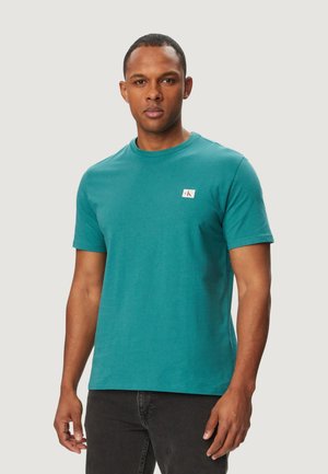 Uomo in piedi rivolto in avanti che indossa una t-shirt a maniche corte color turchese con una piccola toppa con logo sul petto e pantaloni scuri.