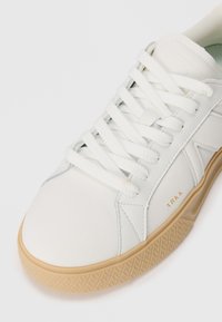 Hvit sneaker med teksturert skinnøvre, flate hvite snørebånd og såle av gummigummi. Har subtil merking i gull på siden.
