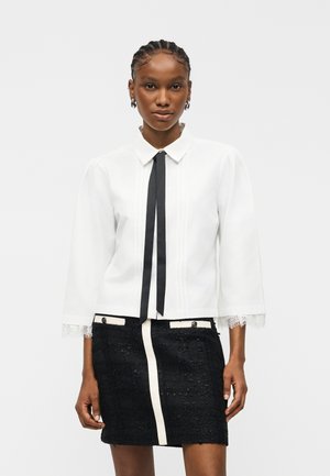 Γυναίκα με πλεγμένα μαλλιά φοράει λευκό μπλουζάκι με μακριά μανίκια, μαύρη κορδέλα δεμένη στον λαιμό και μαύρη υφασμάτινη φούστα με λευκά περιθώρια και λεπτομέρειες με κουμπιά.