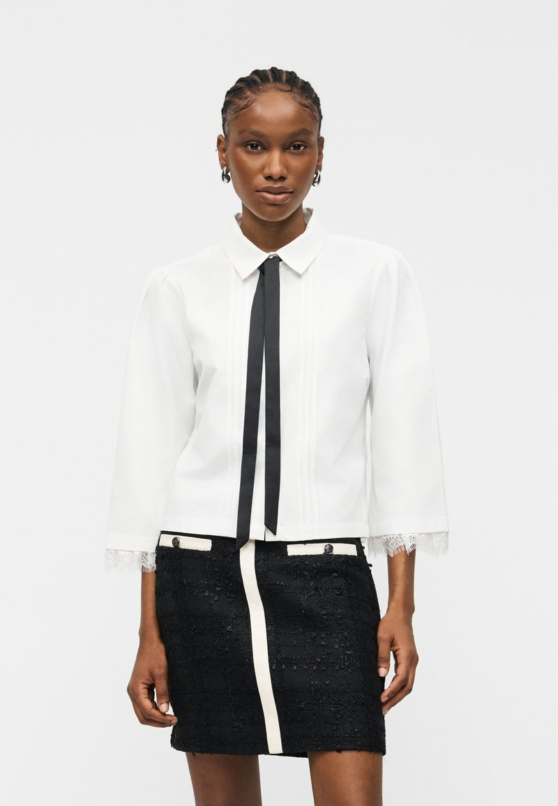 Γυναίκα με πλεγμένα μαλλιά φοράει λευκό μπλουζάκι με μακριά μανίκια, μαύρη κορδέλα δεμένη στον λαιμό και μαύρη υφασμάτινη φούστα με λευκά περιθώρια και λεπτομέρειες με κουμπιά.