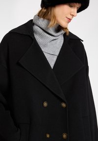 Abrigo negro de doble botonadura en mezcla de lana, con amplias solapas, dos bolsillos frontales y botones en tono dorado, usado sobre un suéter de cuello alto gris.