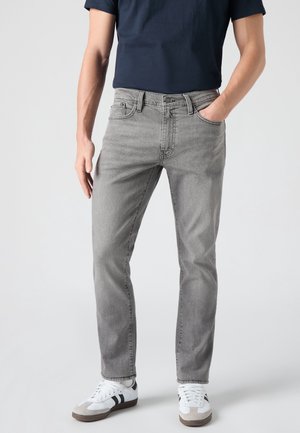 Homme portant un jean gris clair, un t-shirt bleu marine et des baskets blanches avec des bandes noires, debout avec une main dans la poche.