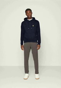 Vīrietis ģērbies jūras zila kapuces džemperī ar mazu dzeltenu logo, pelēkās biksēs un baltās kedās, stāv uz vienkārša gaiša fona.