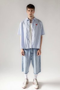 Mlad moški nosi svetlo modro kratkotrajno srajco s kratkimi rokavi in rdečim srčkastim obližem, dolge jeans kratke hlače, bele nogavice in teniske.