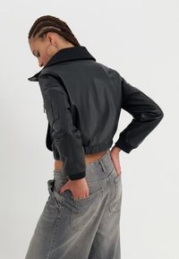 Black faux leather cropped jacket with a large collar, ribbed cuffs, and zippered side pocket. Styled with high-waisted gray jeans.