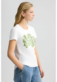 T-shirt en coton blanc avec un motif floral vert et des motifs en spirale sur le devant. Le modèle l'a associé à un jean bleu et des boucles d'oreilles en forme de cœur.