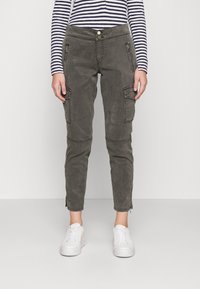 Cargo-Hosen in Olivgrün mit Reißverschlusstaschen, schmalem Schnitt und Knöchelfilzern. Getragen mit weißen Sneakers und einem navy-blauen gestreiften Oberteil.