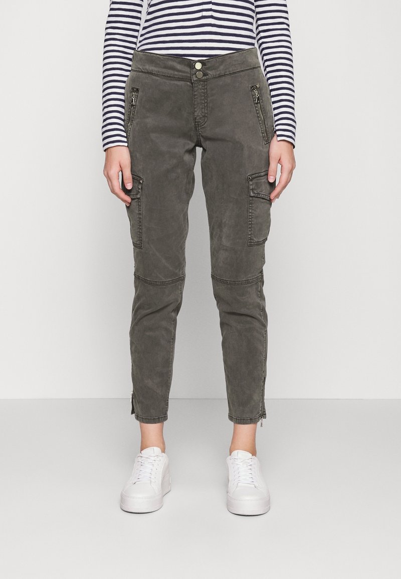 Cargo-Hosen in Olivgrün mit Reißverschlusstaschen, schmalem Schnitt und Knöchelfilzern. Getragen mit weißen Sneakers und einem navy-blauen gestreiften Oberteil.
