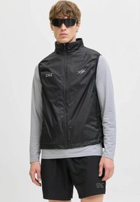 Zwarte mouwloze vest met hoge kraag, voorkant met rits en reflecterende accenten, gedragen over een lichtgrijze lange mouwen shirt en zwarte shorts.