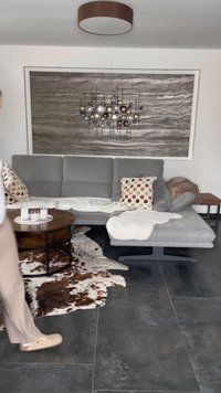 Grå klädd sektionsoffa med mönstrade kuddar, ett runt träkaffebord och en pälsmatta på mörkt kaklat golv. Väggkonst ovanför.