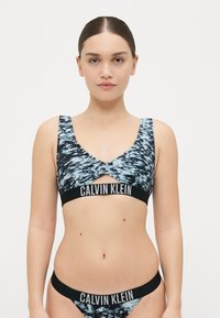 Calvin Klein komplet grudnjaka i bikinija s apstraktnim uzorkom u plavoj i crnoj boji. Mekani materijal, široke naramenice i detalj u vidu logotipa na pojasu.