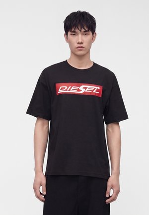 Joven con cabello corto y negro que lleva una camiseta negra de Diesel con un logo rojo y blanco, de pie frente a un fondo liso.
