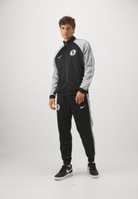 Μαύρο και γκρι αθλητικό κοστούμι με μπουφάν με φερμουάρ, παντελόνια με ελαστική μέση, με λογότυπα και εφαρμοστό σχέδιο. Συνδυασμένο με μαύρα sneakers.