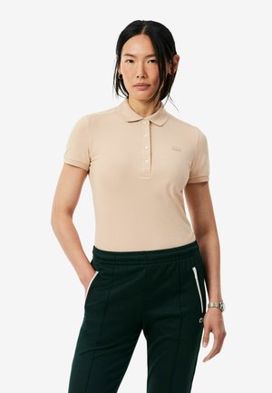 Femme aux cheveux foncés portant un polo beige à manches courtes et un pantalon noir avec des poches zippées blanches, debout, une main dans la poche.