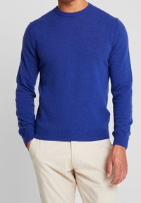 Blå stickad tröja med långa ärmar, rund halsringning och ribbade muddar och nederkant. Bärs med beige byxor som har en slät textur.