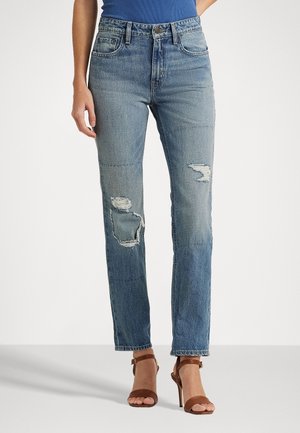 Vrouw draagt blauw mid-rise jeans met versleten plekken op de dijen en bruine sandalen met hoge hakken, staand tegen een eenvoudige achtergrond.