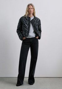 Chaqueta negra acolchada con un cuello alto, combinada con una camisa gris y pantalones anchos negros. Textura suave, diseño minimalista y sin cierres visibles.