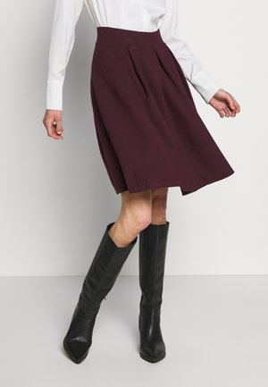 Kvinde iført en knælængde, mørkelilla plisseret nederdel, hvid langærmet bluse og sorte knæhøje læderstøvler.