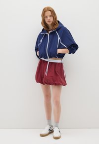 Giovane donna in piedi contro un muro bianco, indossa un giubotto corto con cappuccio di colore blu navy, una gonna a palloncino bordeaux, calze grigie e sneakers bianche.