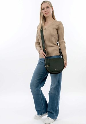 Donna in cardigan a coste beige e jeans a gamba larga tiene in mano una borsa a tracolla nera con sneakers bianche su uno sfondo bianco.